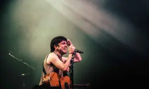 Declan McKenna passionately sings into a microphone, guitar in hand, bathed in stage lights. A glimpse of his setlist is visible.