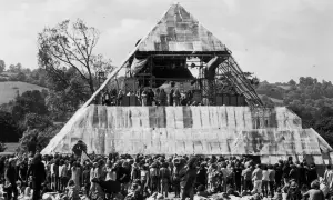 A massive pyramid stage hosts performers at the first Glastonbury Festival. A huge crowd gathers below, marking a historic moment in music history.