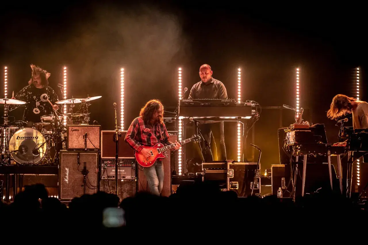The War on Drugs band performs live, bathed in warm stage lights. A guitarist in a red plaid shirt plays center stage, with a drummer, keyboardist, and another musician on synths.