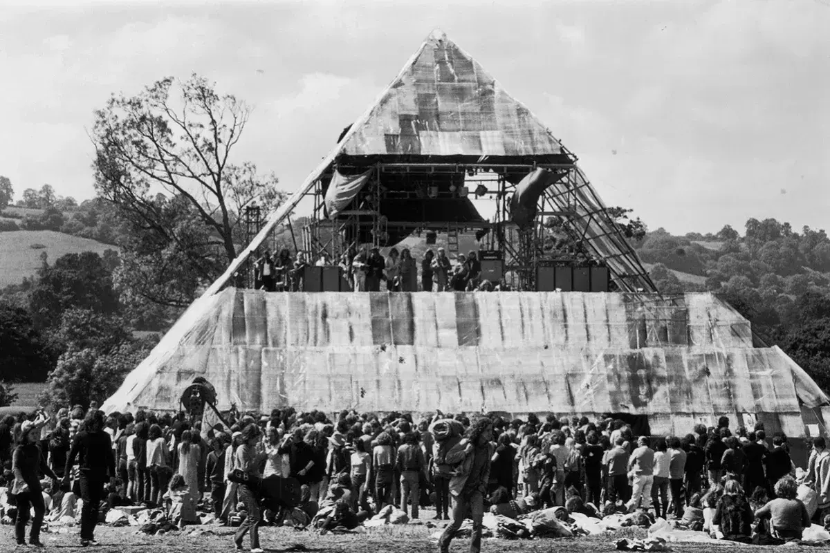 A massive pyramid stage hosts performers at the first Glastonbury Festival. A huge crowd gathers below, marking a historic moment in music history.