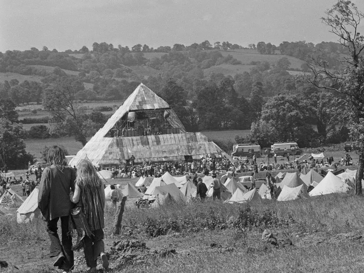 A couple walks towards the iconic pyramid stage at the first Glastonbury Festival. Tents and vans dot the landscape.
