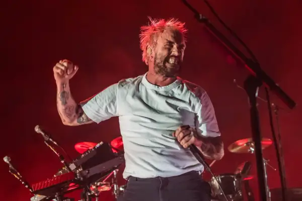 A tattooed singer with spiky blonde hair belts into a microphone, fist raised, during a live performance. The idles setlist is in full swing.