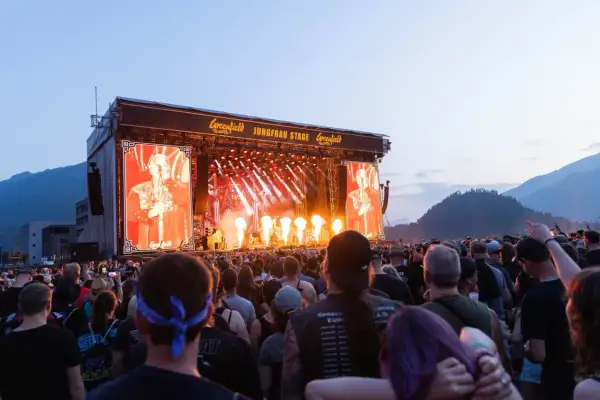 A massive crowd enjoys a performance at the Greenfield festival, with a large screen displaying a performer and flames erupting on stage.