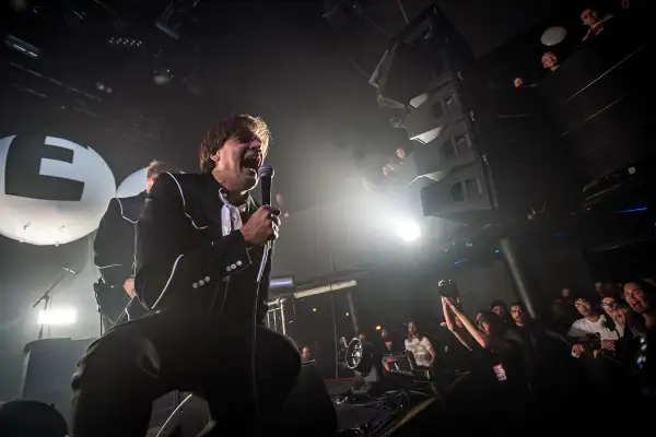 The Hives setlist is being performed by a charismatic frontman in a black suit, belting into a microphone on a smoky stage. The crowd is visible, enjoying the energetic show.