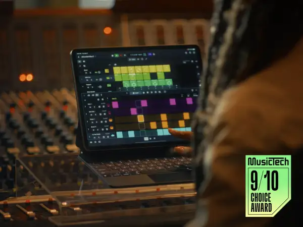 A person's finger touches a tablet displaying Logic Pro for iPad, showing a colorful grid of musical patterns, in a recording studio.